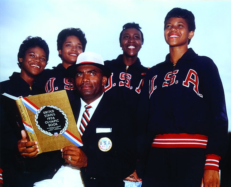 Coach Ed Temple and the U.S. Women's team look off camera. Coach Temple holds the United States 1956 Olympic Book. Track star Wilma Rudolph stands on the far right.