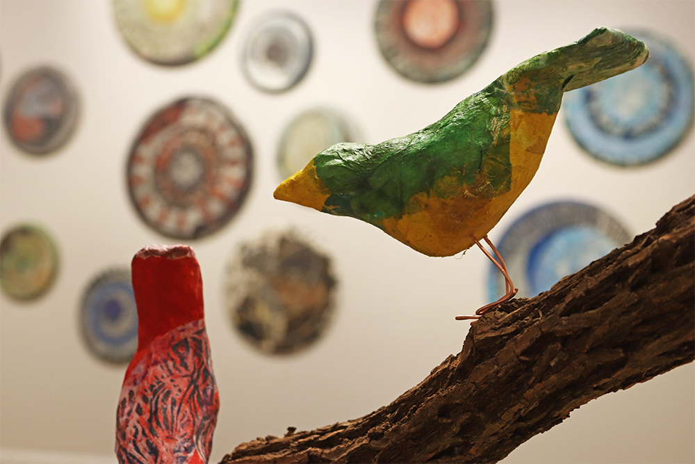 Papier-mâché birds sitting on a branch. Large decorative circular pieces are hanging on the wall in the background.