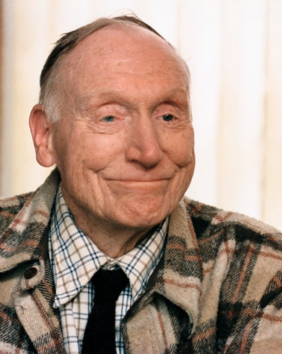 Robert Penn Warren wearing a flannel coat over a plaid button up and dark tie. He's smiling softly off camera.