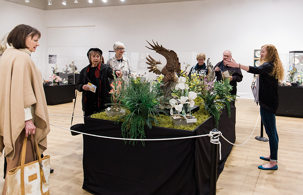 A group of older visitors peer at a Boehm porcelain display while a young tour guide motions towards the porcelain pieces.